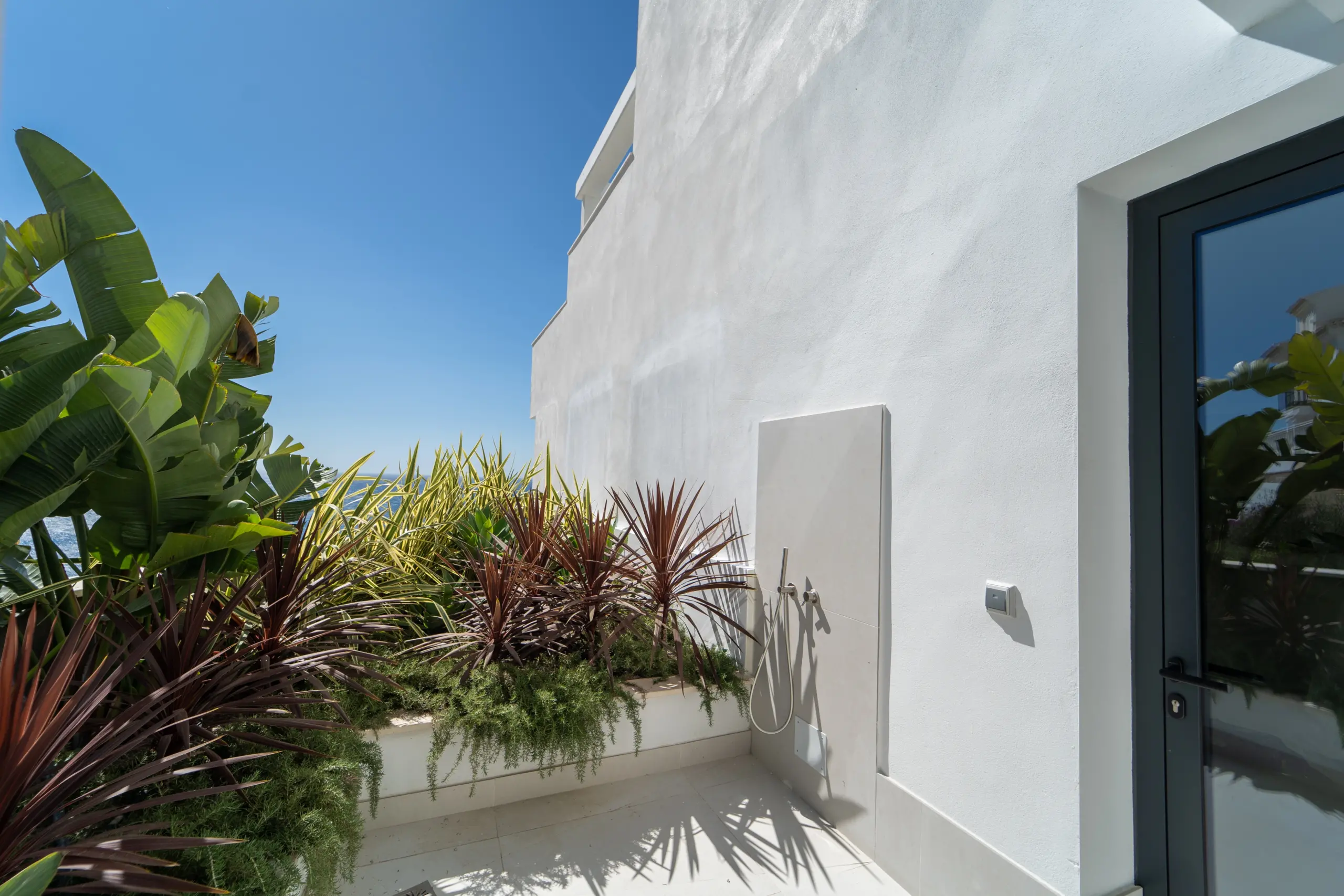 Imagen de una ducha exterior situada junto a un muro blanco liso, con una instalación moderna de metal. Rodeada por un exuberante jardín tropical que incluye grandes hojas verdes de platanero y plantas de follaje rojo y amarillo, creando un ambiente natural y relajante. El cielo despejado y soleado ofrece una sensación de frescura y tranquilidad.