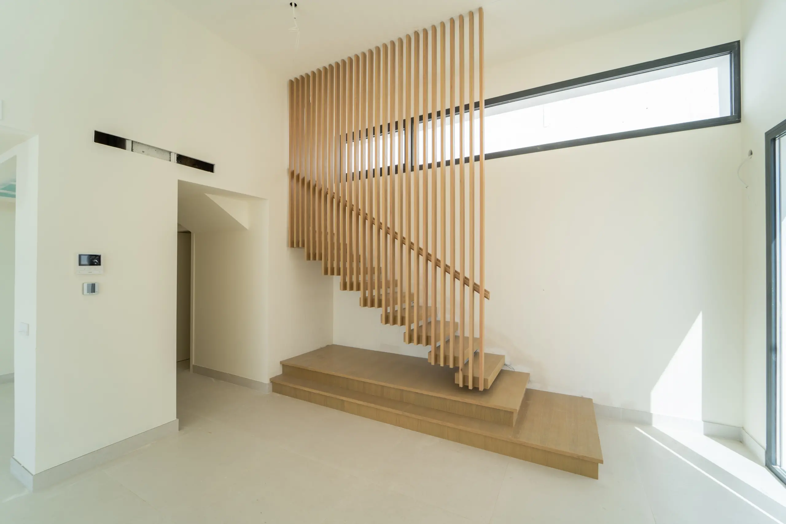 Imagen de una escalera de madera con barandilla de listones verticales, ubicada en un espacio amplio y bien iluminado. La escalera, de tonos madera clara, cuenta con una estructura de escalones anchos que se integran armónicamente en un entorno de paredes blancas. Un ventanal largo y estrecho en la parte superior permite la entrada de luz natural, destacando la simplicidad y elegancia del diseño.