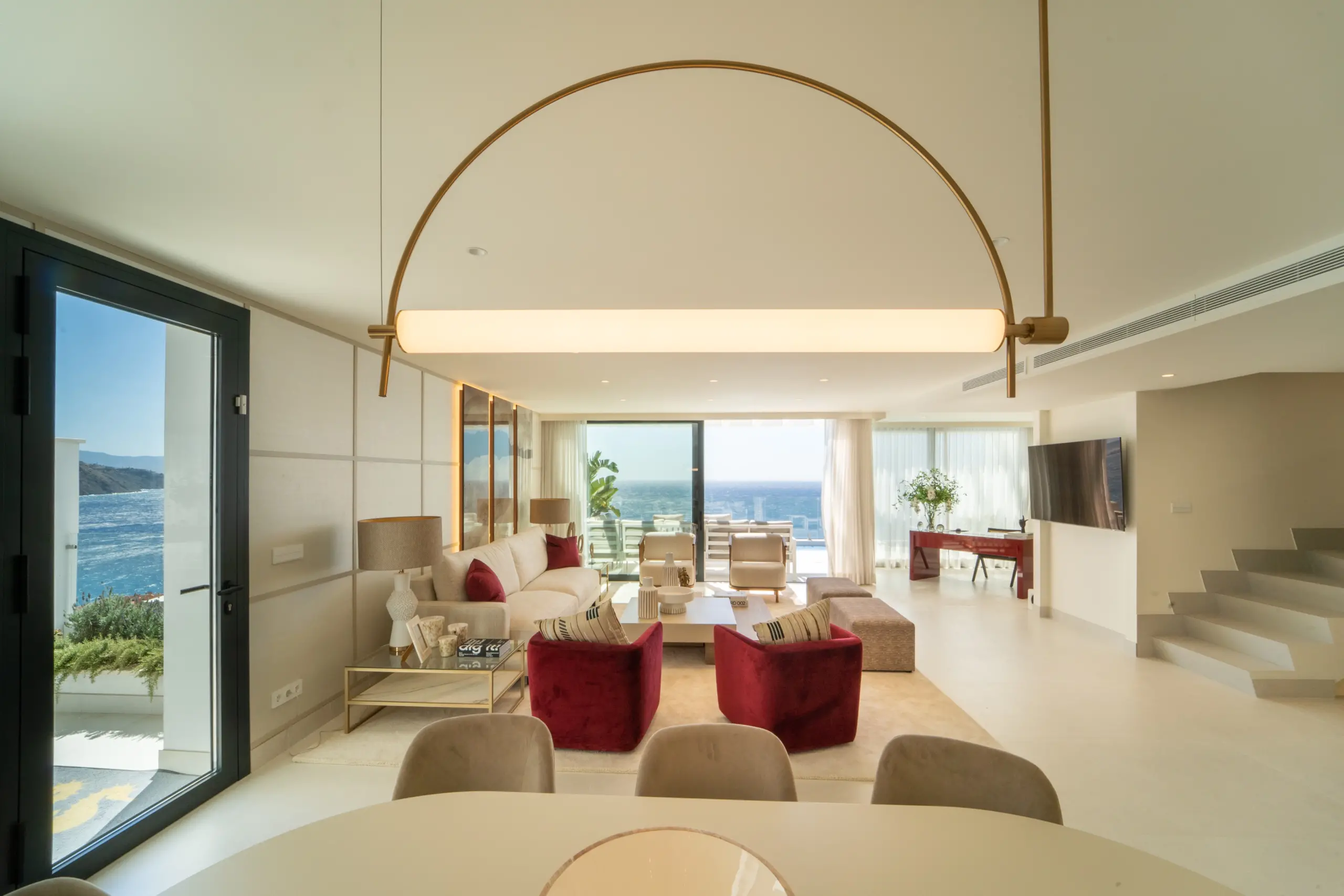 Espacio abierto y luminoso que combina salón y comedor. El área del salón incluye sofás de tonos neutros y rojo, mientras que el comedor tiene una mesa redonda grande rodeada de sillas de terciopelo. Decoración moderna con toques de diseño contemporáneo y arte moderno.