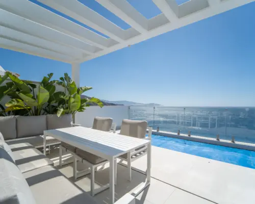 Espaciosa terraza con techo de pérgola blanca, equipada con un sofá largo de color gris claro y una mesa de comedor blanca con sillas a juego. La terraza ofrece una vista impresionante del mar Mediterráneo, rodeada de exuberante vegetación tropical. Una piscina visible al fondo añade un toque relajante y lujoso al espacio exterior.
